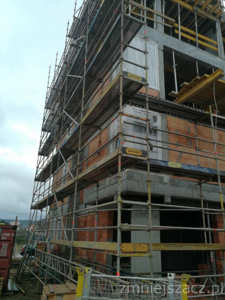 Wznoszony budynek z cegły i betonu otoczony rusztowaniem z widocznym kontenerem na pierwszym planie.