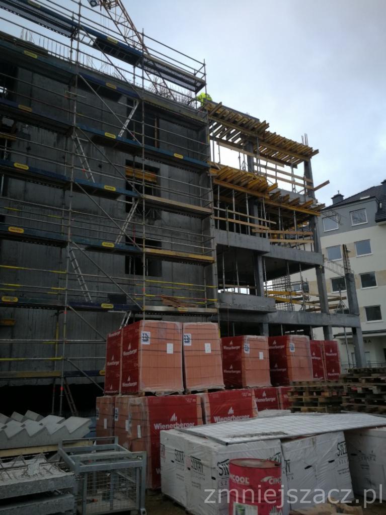 Wznoszony budynek z betonu i cegły, z widocznymi rusztowaniami, szalunkami i paletami cegieł Wienerberger na pierwszym planie.