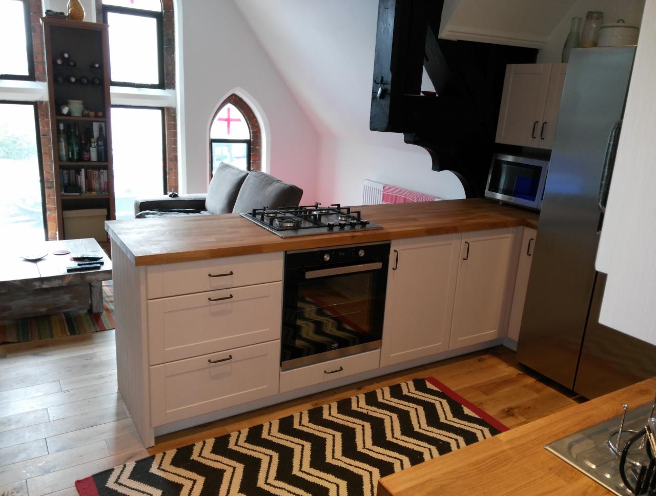 Wnętrze kuchni z białymi szafkami, drewnianym blatem, czarną kuchenką i piekarnikiem, oraz stalową lodówką. W tle salon z oknem w kształcie łuku i ceglaną ścianą.