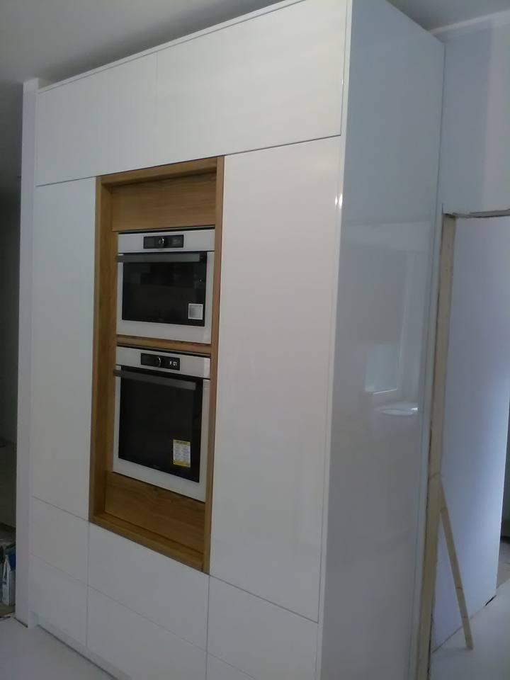 Biała, lakierowana zabudowa kuchenna z dwoma piekarnikami w drewnianej obudowie, widoczna z boku, z elementami wykończeniowymi w tle.