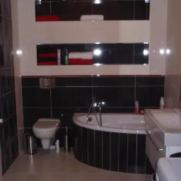 Wykończona łazienka z czarno-białymi płytkami, wanną narożną, umywalką i toaletą. Na ścianie wnęki z ręcznikami. Grzejnik drabinkowy po lewej stronie.