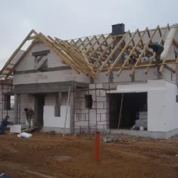 Budowa domu jednorodzinnego: murowane ściany z bloczków, drewniana konstrukcja dachu, ocieplenie styropianem. Prace budowlane w toku, widoczni robotnicy i rusztowania.