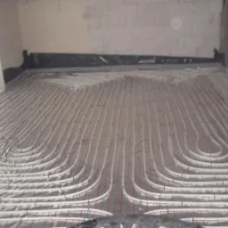 Białe rurki ogrzewania podłogowego ułożone na srebrnej folii, widoczne szare ściany z betonu komórkowego i okno po lewej stronie.