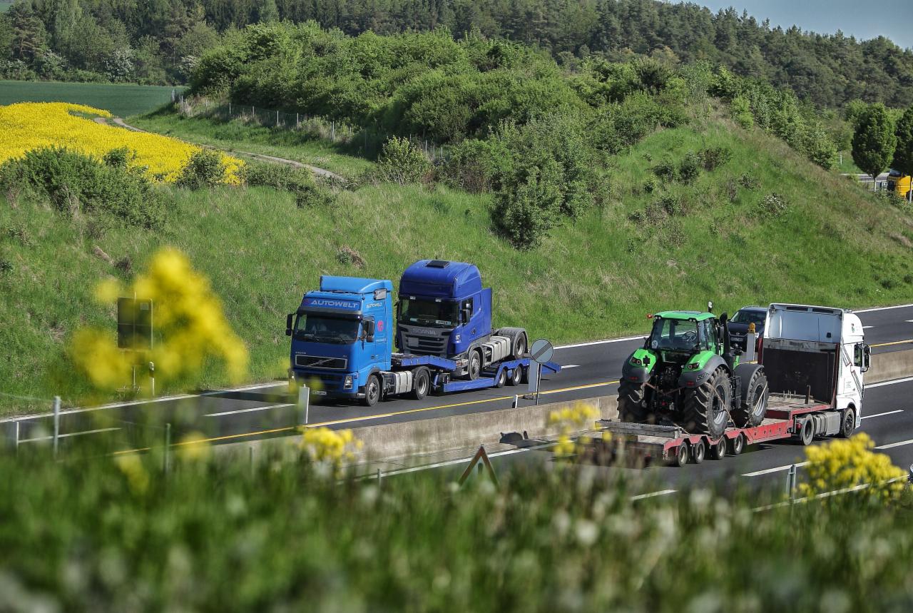 Transport ciężarowy: Dwie ciężarówki, jedna niebieska z napisem Autowelt, przewożąca drugą, ciemnoniebieską ciężarówkę i zielony traktor na niskopodwoziowej naczepie, jadące po autostradzie...