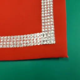 Fragment czerwonej tkaniny z dekoracyjnym, srebrnym, błyszczącym obramowaniem, leżącej na zielonej powierzchni.