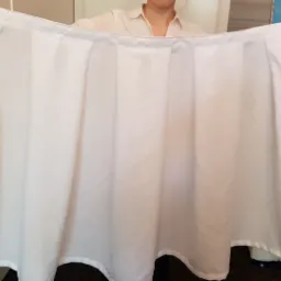 Biała, plisowana tkanina z wykończonym brzegiem, trzymana przez osobę w jasnej koszuli na tle ściany i sofy.