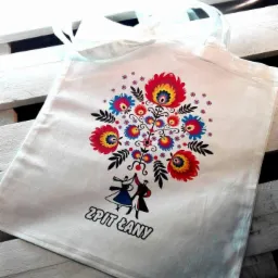 Biała torba z nadrukiem ludowym kwiatów i tańczącej pary w strojach ludowych, leżąca na białym, drewnianym tle.