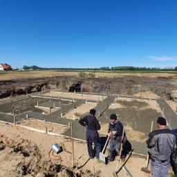 Przedsiębiorstwo Budowlano Transportowe Stefan Zakrzewski - Zalane betonem szalunki fundamentowe pod budowę domu jednorodzinnego, widoczni pracownicy budowlani i wykopany teren pod fundament, w tle krajobraz z domem.