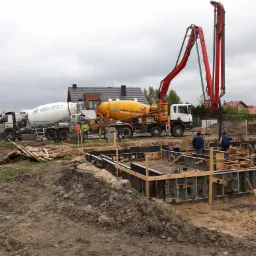 Dwa betonomieszarki i pompa do betonu na placu budowy domu jednorodzinnego; widoczne szalunki fundamentowe i pracownicy w trakcie wylewania betonu.