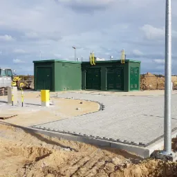 Nowo wybrukowany plac z kostki brukowej z elementami infrastruktury gazowej i dwoma zielonymi budynkami transformatorowymi w tle, widoczny także żółty traktor z paletą kostki.