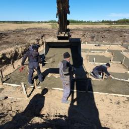 Przedsiębiorstwo Budowlano Transportowe Stefan Zakrzewski - Budowa fundamentów domu jednorodzinnego: robotnicy wyrównują wylany beton w szalunkach, z łyżki koparki wysypuje się kolejna porcja mieszanki.