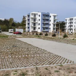 Nowo wybudowany parking z ażurowych płyt betonowych przed nowoczesnym budynkiem apartamentowym, z widoczną koparką i przyczepą kempingową w tle.