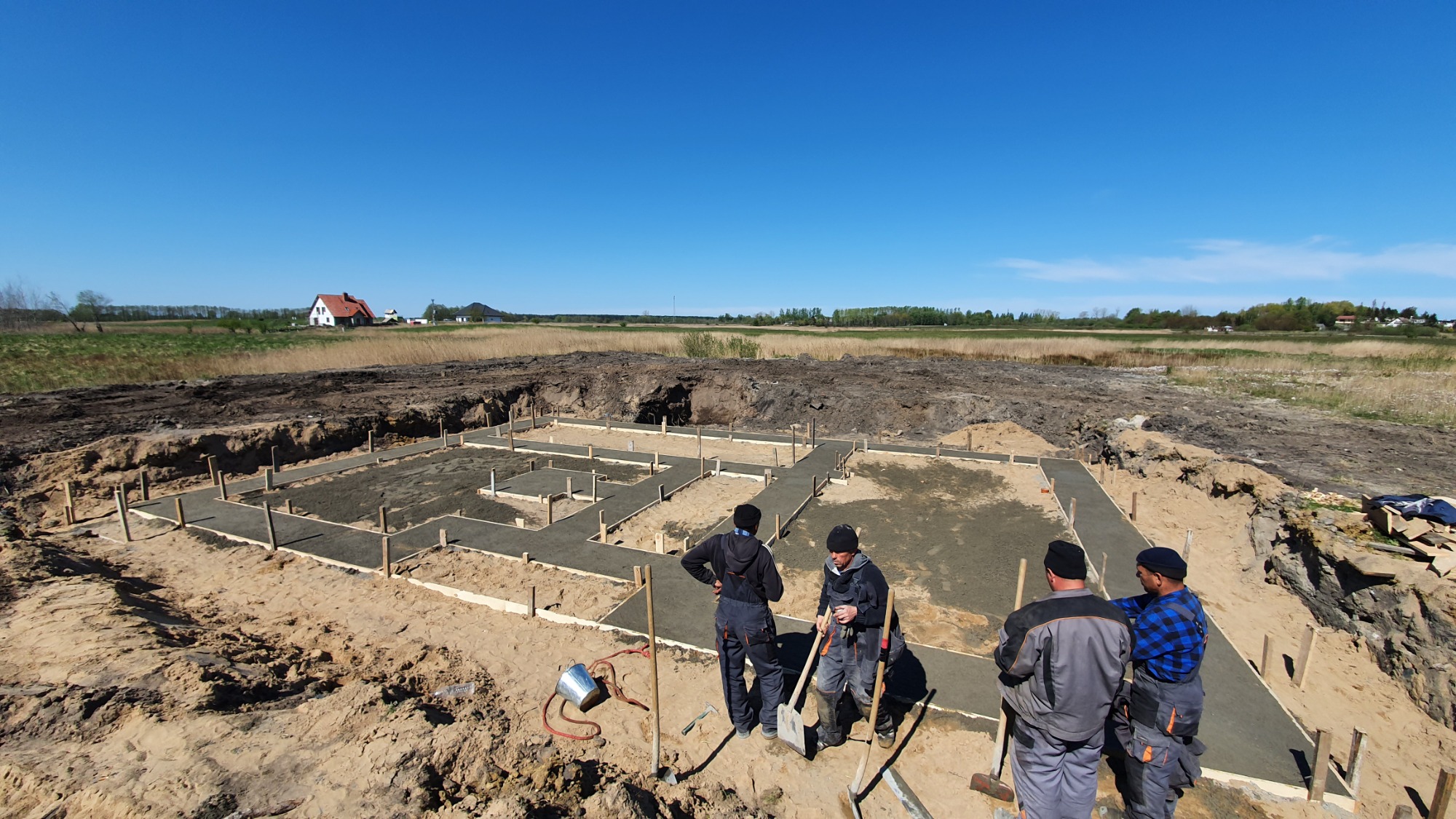 Zalane betonem szalunki fundamentowe pod budowę domu jednorodzinnego, widoczni pracownicy budowlani i wykopany teren pod fundament, w tle krajobraz z domem.
