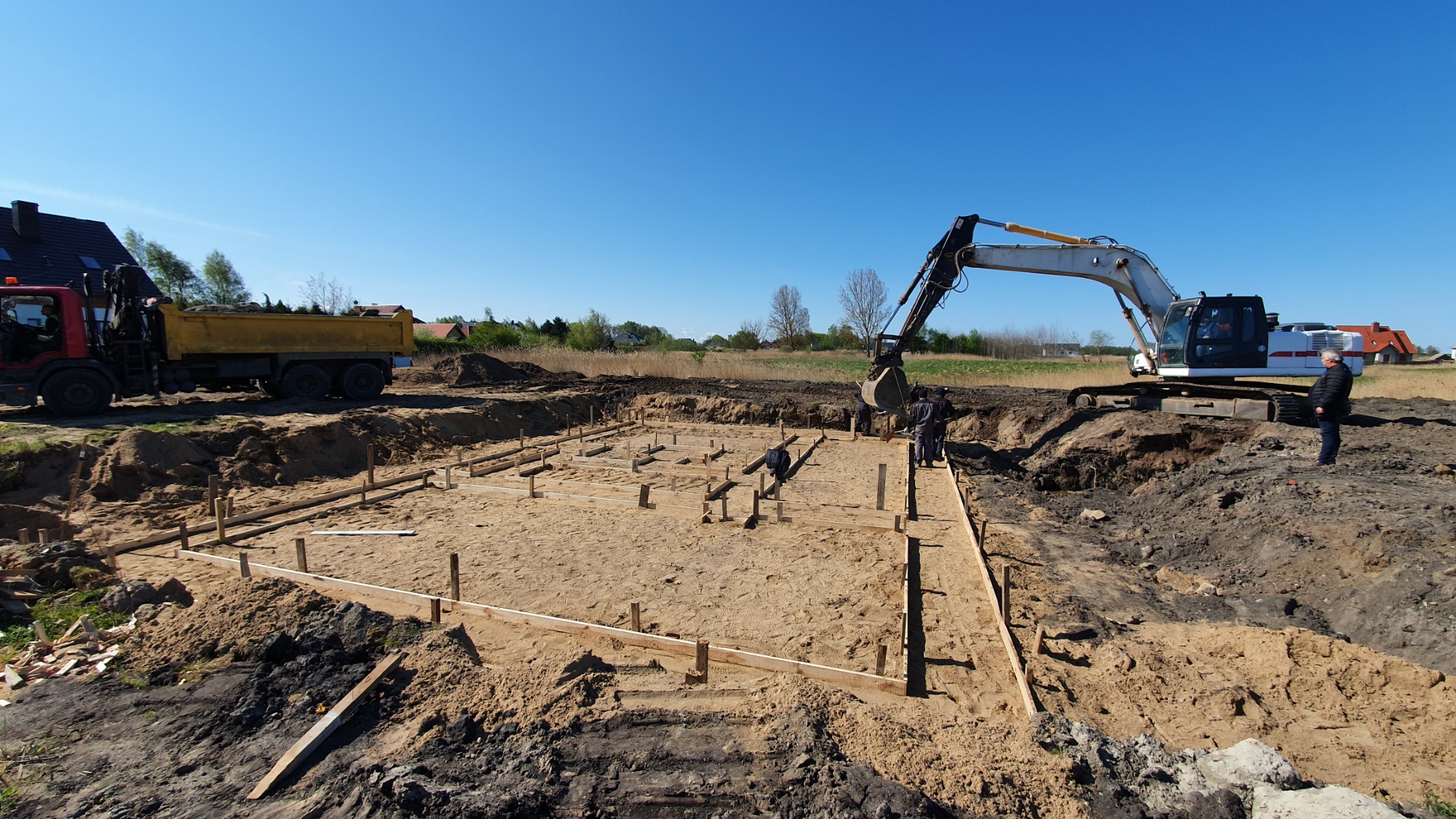 Przygotowanie terenu pod budowę domu jednorodzinnego: wykop z drewnianym szalunkiem, ciężarówka i koparka w tle, pracownicy przy wykopie.