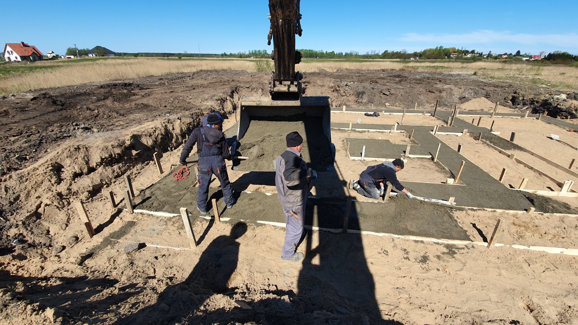Budowa fundamentów domu jednorodzinnego: robotnicy wyrównują wylany beton w szalunkach, z łyżki koparki wysypuje się kolejna porcja mieszanki.
