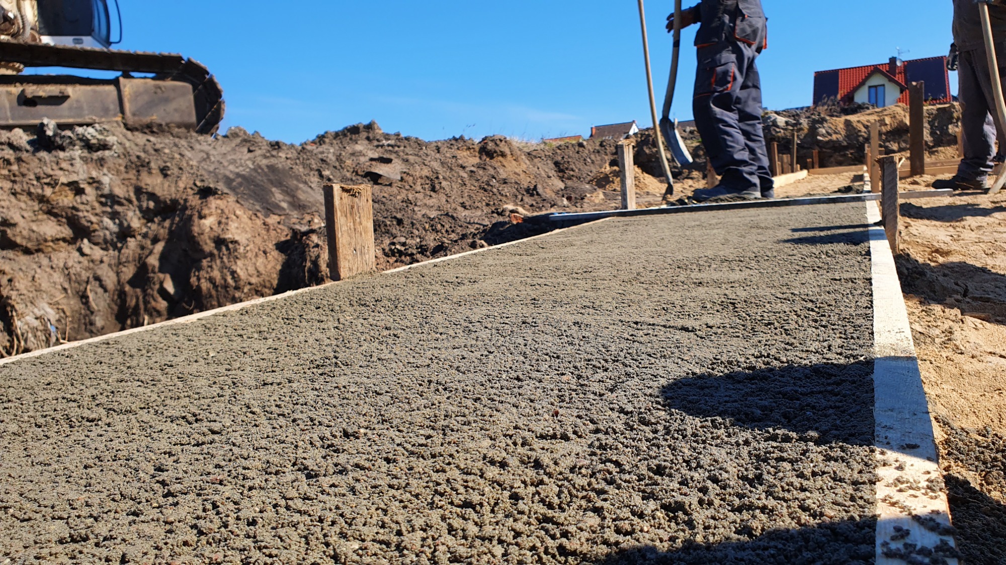 Wylany beton na ławę fundamentową pod dom jednorodzinny, widoczne drewniane szalunki, wykop ziemny w tle i fragment koparki.