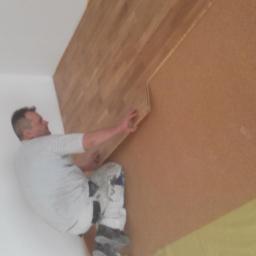 Usługi Ogólnobudowlane Adam Bednarski - Mężczyzna w ubrudzonych farbą spodniach układa drewniane panele na ścianie pokrytej korkiem w trakcie prac wykończeniowych.