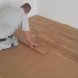 Usługi Ogólnobudowlane Adam Bednarski - Układanie drewnianej podłogi: mężczyzna na kolanach dopasowuje kolejną deskę na podkładzie korkowym, widoczne narzędzia pomiarowe i kliny dystansowe.