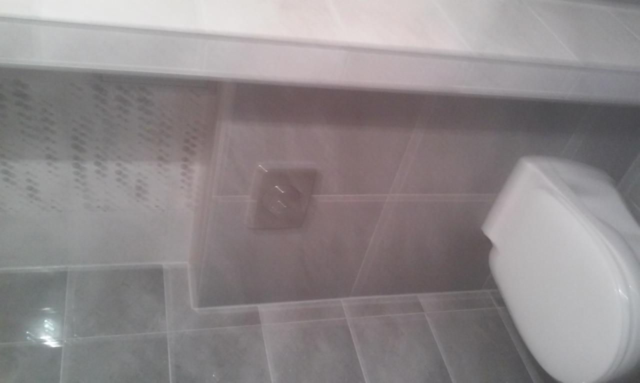 Szare płytki w łazience, widoczny fragment toalety, gniazdko elektryczne na ścianie i wzorzysty sufit.