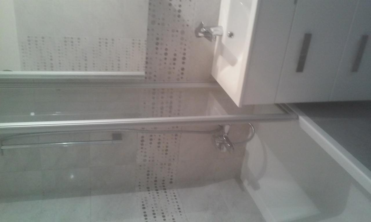 Nowoczesna łazienka z kabiną prysznicową z przesuwnymi drzwiami, umywalką z szafką i baterią, wykończona płytkami w odcieniach szarości z delikatnym wzorem.