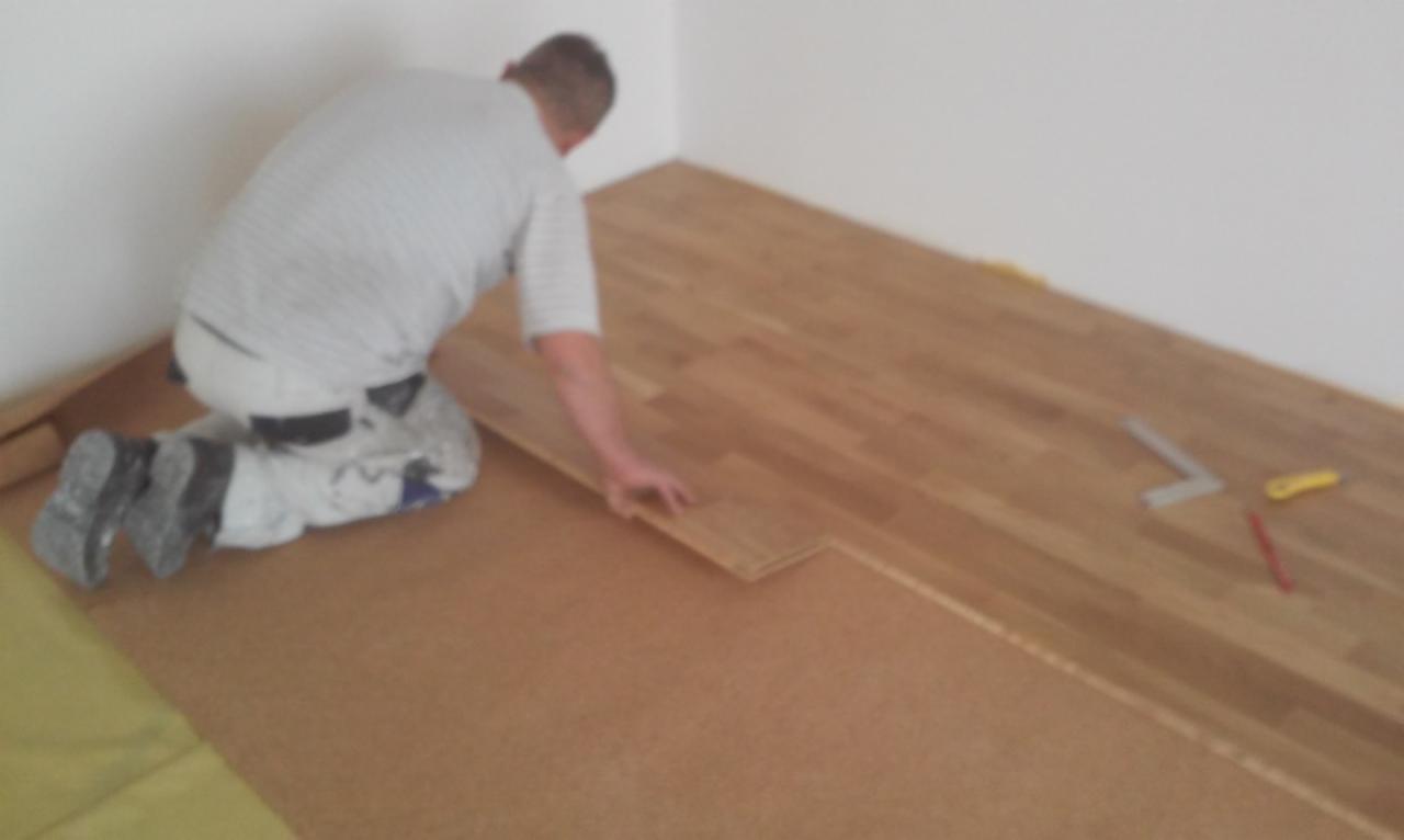 Układanie drewnianej podłogi: mężczyzna na kolanach dopasowuje kolejną deskę na podkładzie korkowym, widoczne narzędzia pomiarowe i kliny dystansowe.