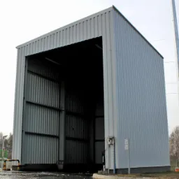 Szara hala z blachy falistej z otwartym wjazdem, widoczna instalacja gazowa i słup energetyczny w tle, pochmurny dzień.