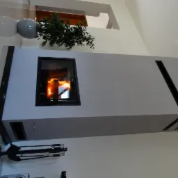 Nowoczesny kominek w zabudowie z szarego betonu architektonicznego, z czarnym wkładem kominkowym i zestawem narzędzi kominkowych obok, uzupełniony rośliną doniczkową.