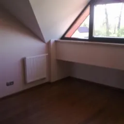 Pokój na poddaszu z drewnianą podłogą, grzejnikiem i oknem w kształcie trójkąta. Ściany pomalowane na jasny kolor, widoczne gniazdka elektryczne.