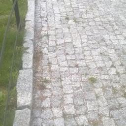 Ka-Bruk - Szara ścieżka z kostki granitowej z widocznymi spoinami i drobną roślinnością, obrzeża ścieżki z większych kamieni, w tle trawa i fragment metalowego ogrodzenia.