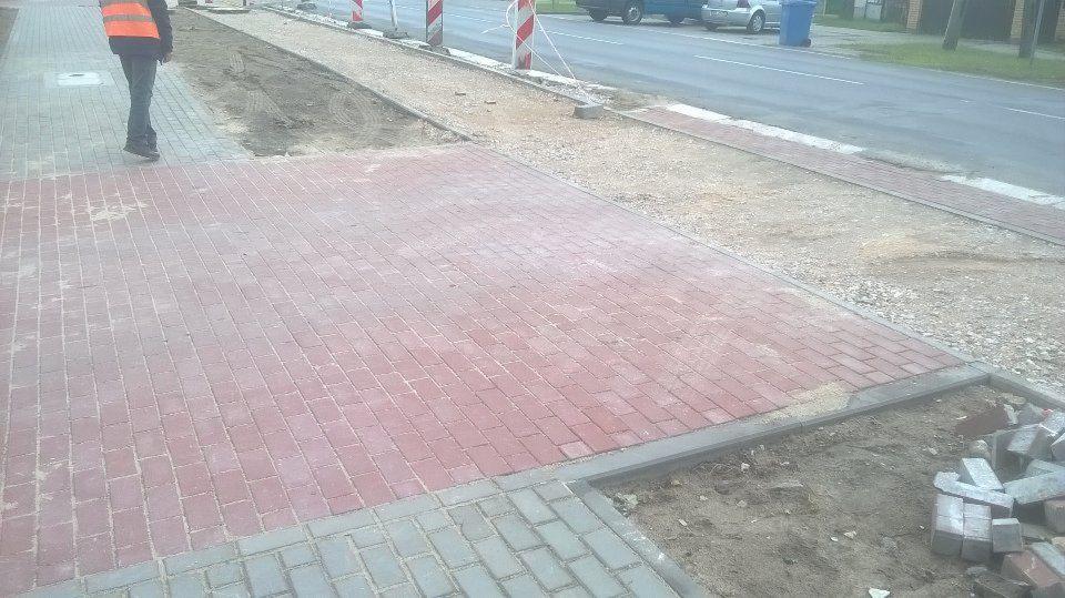 Częściowo ułożona czerwona kostka brukowa na chodniku, widoczny pracownik w pomarańczowej kamizelce, krawężniki i przygotowane podłoże z piasku.