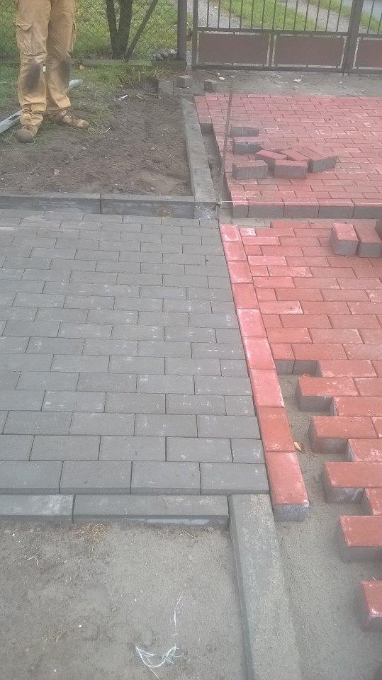 Fragment układanej nawierzchni z kostki brukowej w dwóch kolorach: szarym i czerwonym, widoczne rozłożone kostki i częściowo ułożona szara kostka, obrzeża krawężnikowe, w tle ogrodzenie z siatki.