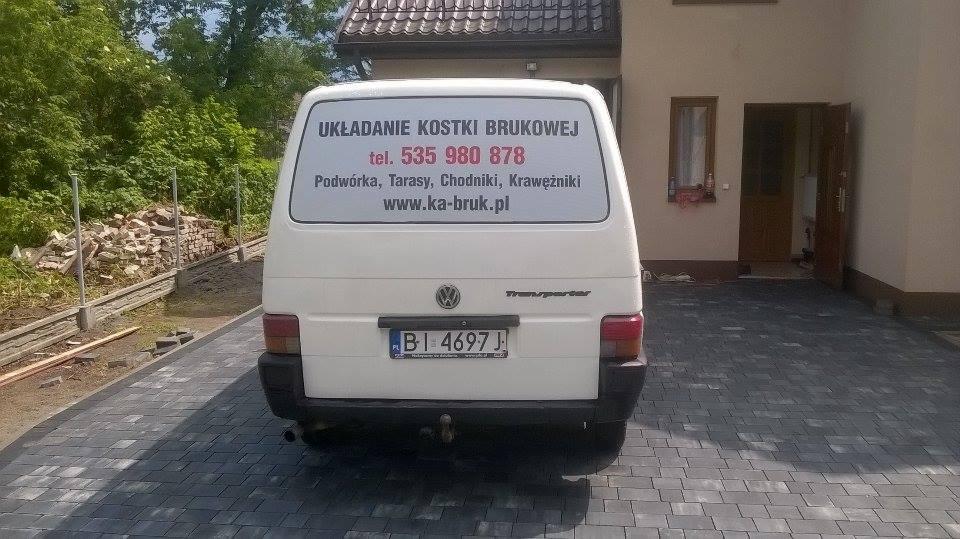 Biały samochód dostawczy z reklamą na tylnej szybie, informującą o usługach układania kostki brukowej, zaparkowany na nowo ułożonym podjeździe z kostki brukowej w odcieniach szarości. W tle sterta...