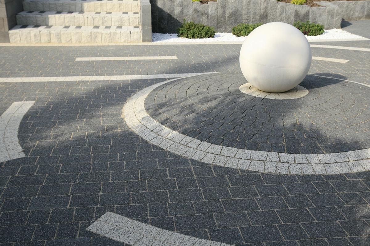 Aranżacja przestrzeni z ciemnej kostki brukowej, dekoracyjna biała kula, schody z kostki i rabata. Estetyczny i nowoczesny wygląd podjazdu.