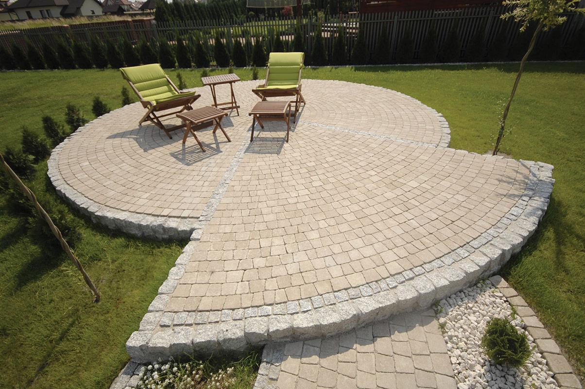 Okrągły taras z jasnej kostki brukowej, obrzeża z szarego granitu. Na tarasie drewniane krzesła i stoliki, w tle trawnik i ogrodzenie z tujami.