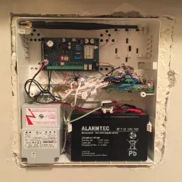 Otwarta obudowa centrali alarmowej z widocznymi płytkami drukowanymi, akumulatorem Alarmtec i transformatorem Noratel. Widoczne okablowanie i antena.