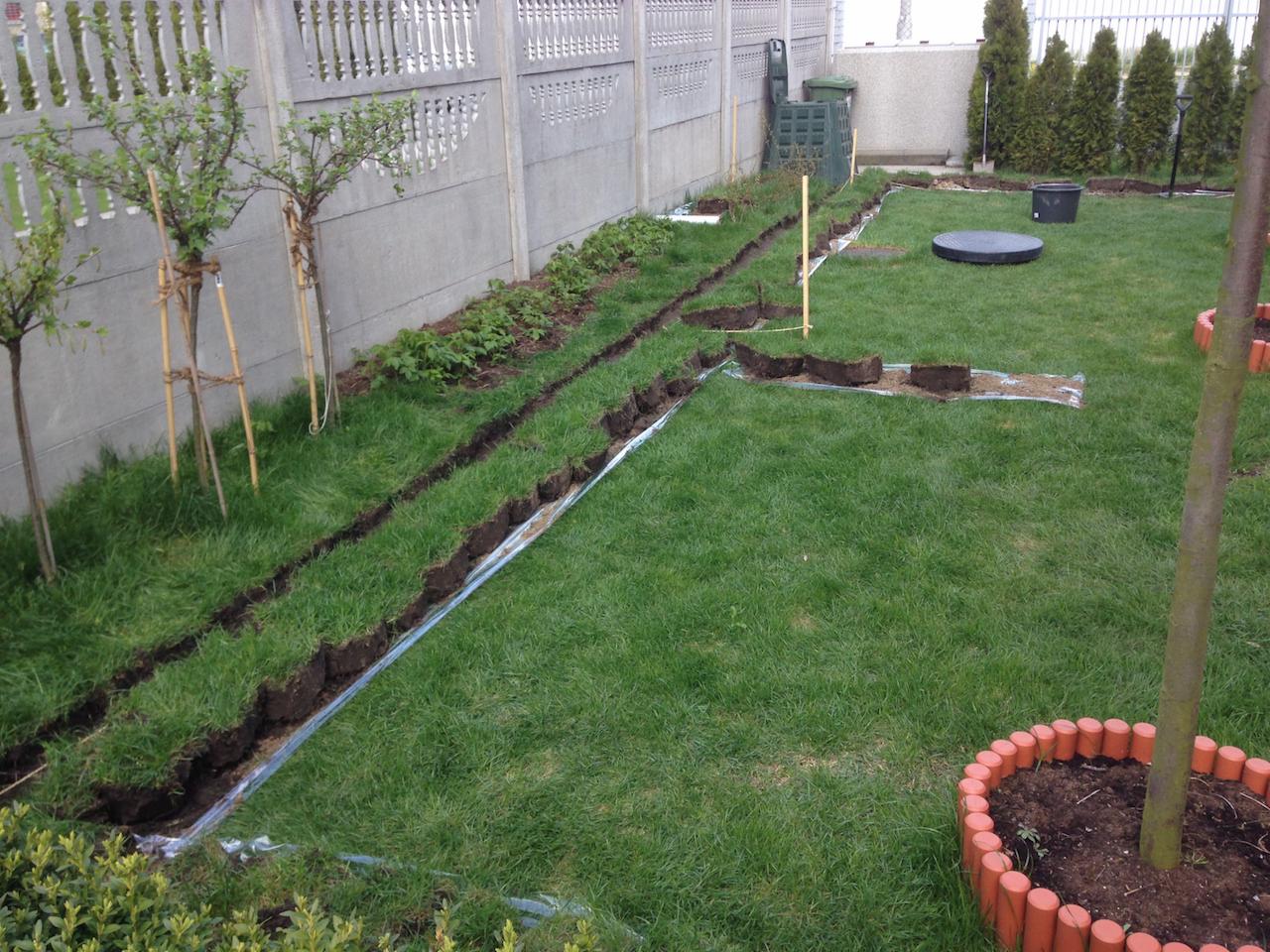 Wykopane rowy w trawniku, zabezpieczone folią, prawdopodobnie pod instalację elektryczną w ogrodzie. Widoczny betonowy płot i drzewa.