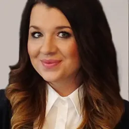 Portret uśmiechniętej kobiety o długich, ombre włosach, ubrana w białą koszulę i ciemną marynarkę, na jasnym tle.