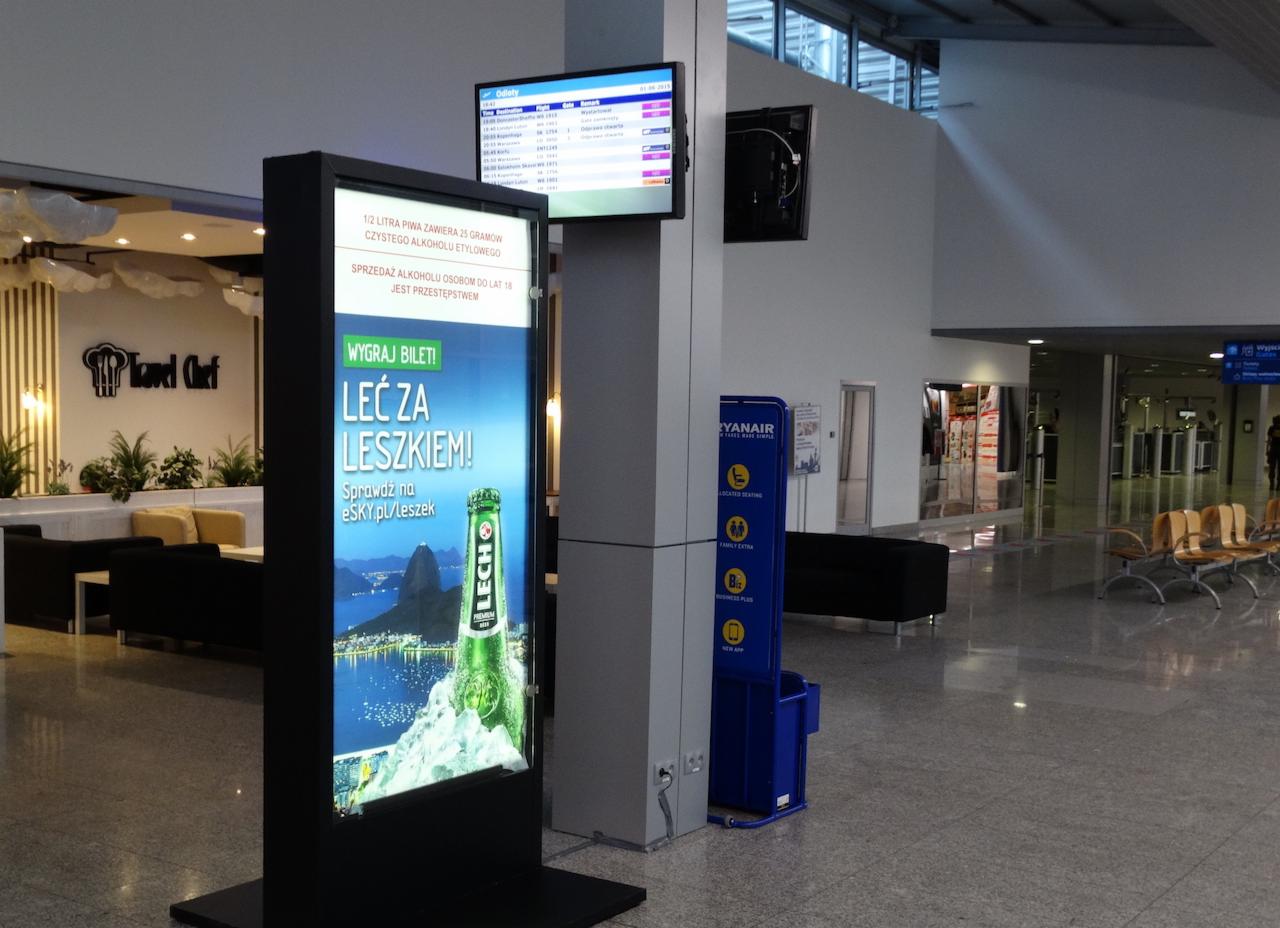 Billboard z reklamą piwa Lech w hali lotniska, obok tablica z odlotami i punkt informacyjny Ryanair, w tle widoczne logo restauracji Travel Chef.