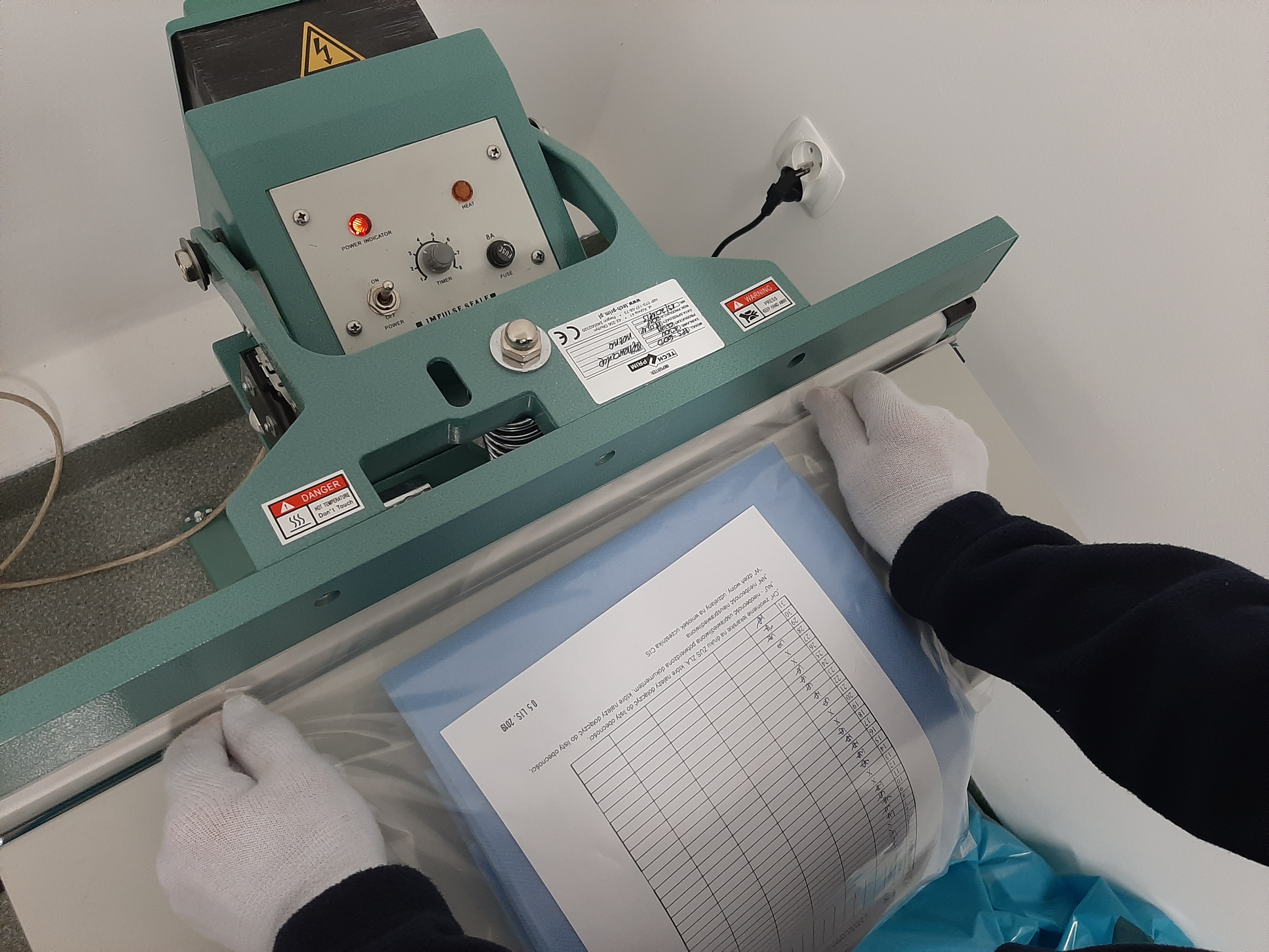 Operator w białych rękawicach umieszcza dokument w niebieskiej folii w zgrzewarce impulsowej z widocznymi włącznikami, pokrętłem timera i lampkami kontrolnymi.