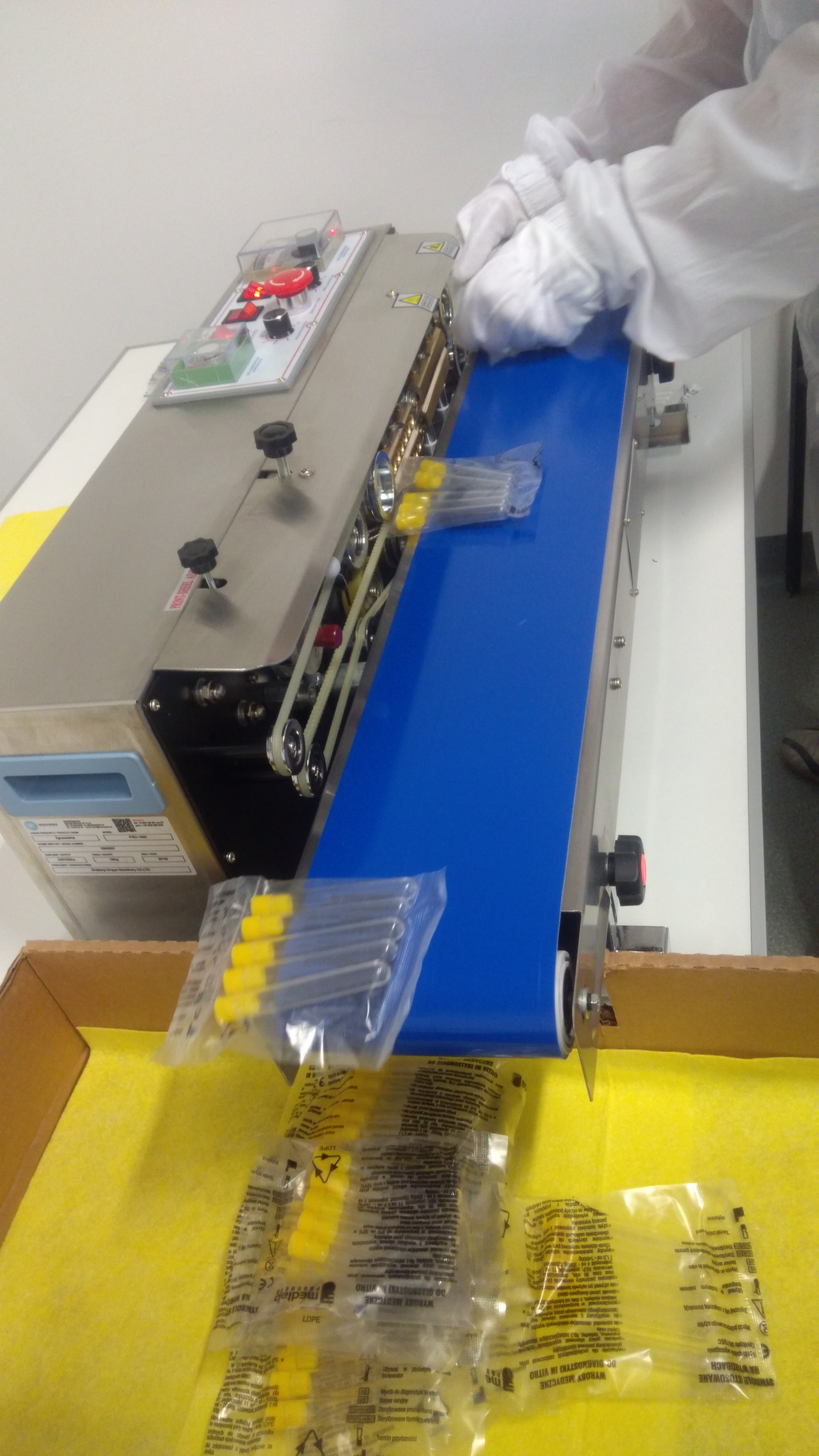 Automatyczna linia do pakowania zestawów medycznych z żółtymi elementami, operator w białym kombinezonie umieszcza zapakowane produkty na niebieskim taśmociągu, karton z zapakowanymi produktami w tle.