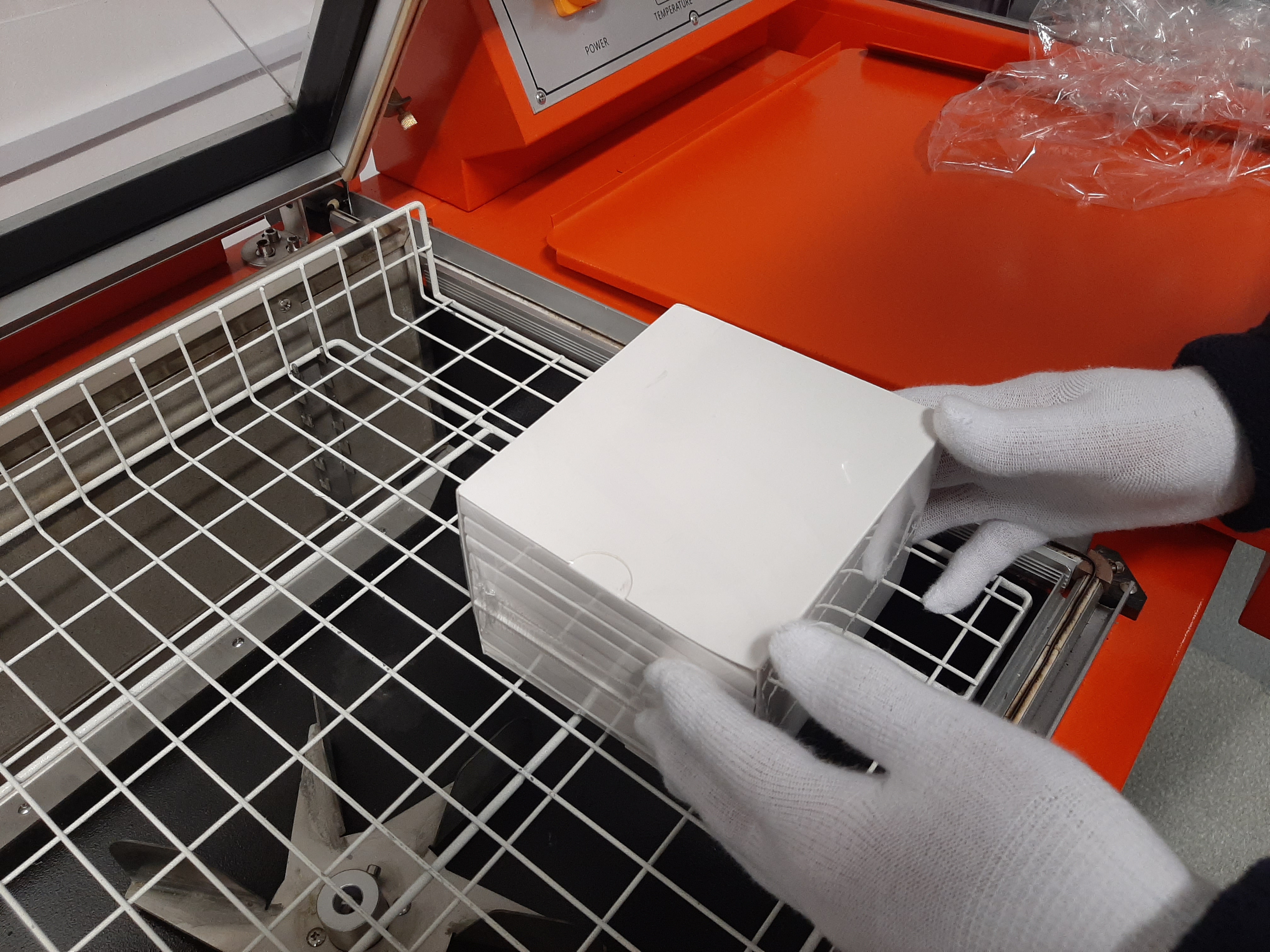 Białe, kwadratowe pudełka układane w stos wewnątrz maszyny do pakowania termokurczliwego przez osobę w białych rękawiczkach, widoczna metalowa kratka maszyny i pomarańczowa obudowa.