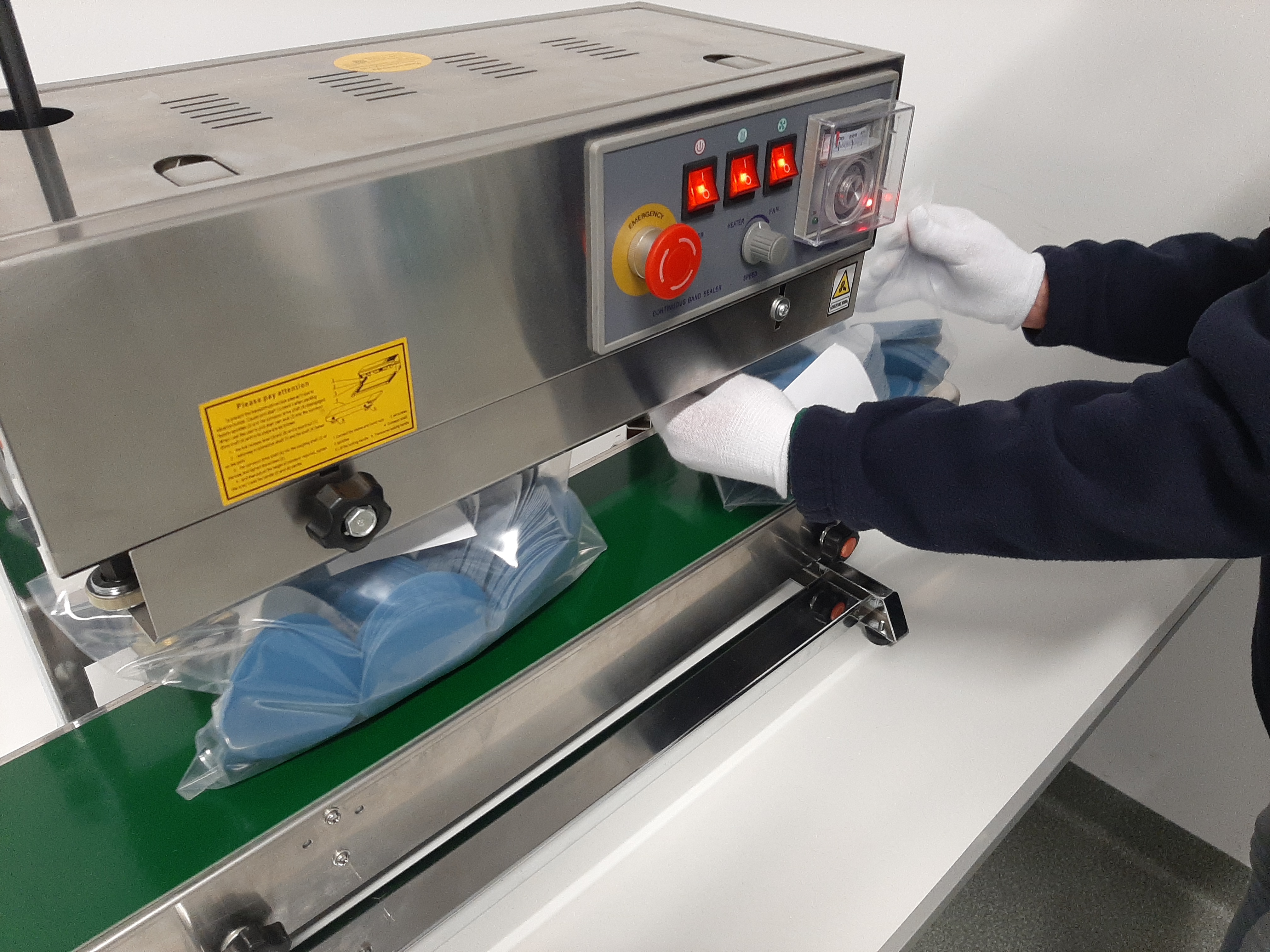 Automatyczna zgrzewarka foliowa z zielonym taśmociągiem, pakująca niebieskie przedmioty w plastikowe worki, obsługiwana przez osobę w białych rękawiczkach.