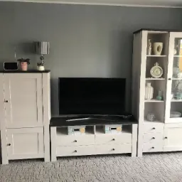 Zestaw mebli do salonu w stylu skandynawskim: białe szafki z ciemnymi blatami, szklana witryna z dekoracjami, telewizor na niskiej komodzie z szufladami i koszykami.