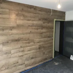 Ściana wyłożona panelami imitującymi drewno, widoczne otwory na gniazdka elektryczne i zabezpieczona folią podłoga w trakcie remontu.