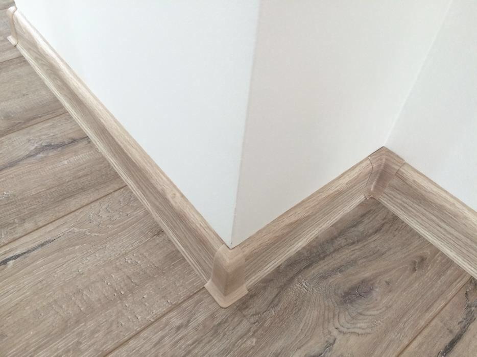 Jasne panele podłogowe z drewnopodobnym wzorem, listwy przypodłogowe w kolorze paneli, narożnik listwy przypodłogowej, białe ściany w rogu pomieszczenia.