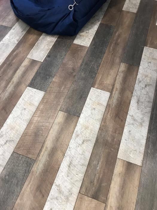 Ułożone pod kątem panele podłogowe w odcieniach brązu, szarości i bieli, z widocznym fragmentem granatowej pufy w rogu.