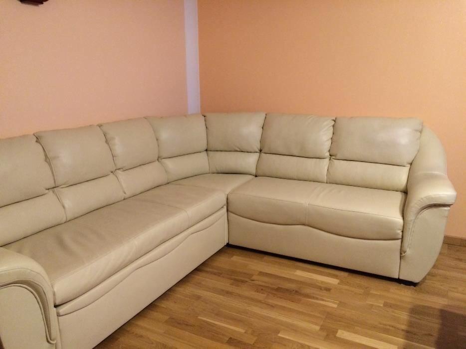 Jasny, skórzany narożnik z wyraźnie widocznym łączeniem elementów siedziska, ustawiony na podłodze z paneli w odcieniach brązu, na tle ściany w kolorze brzoskwiniowym.