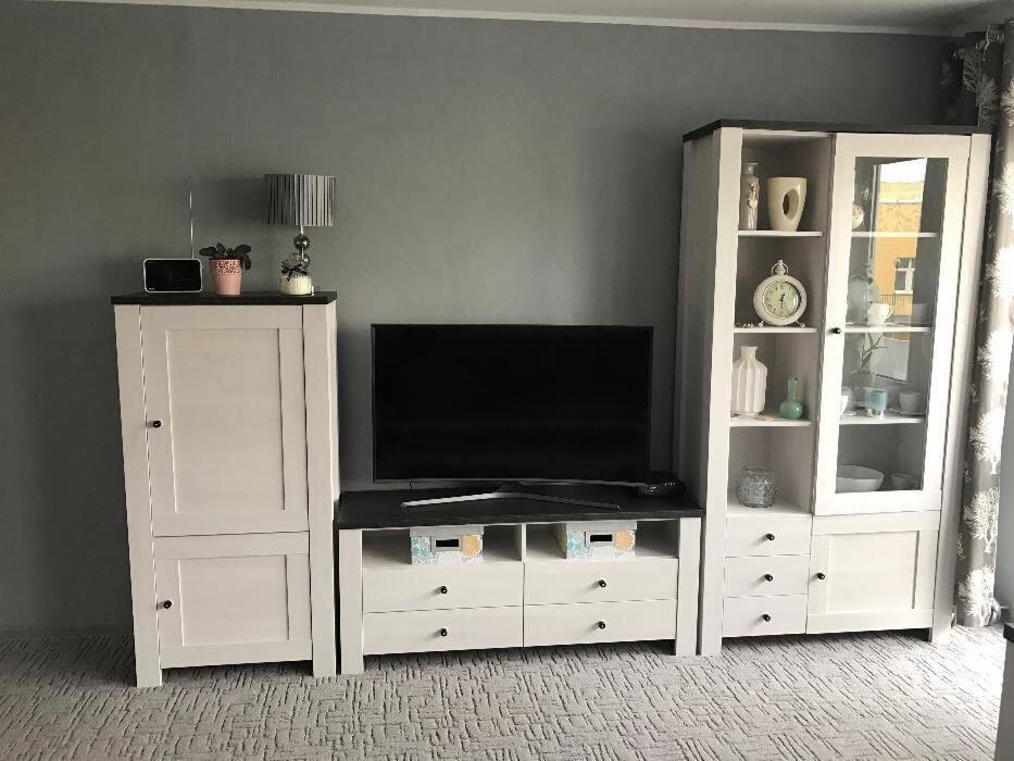 Zestaw mebli do salonu w stylu skandynawskim: białe szafki z ciemnymi blatami, szklana witryna z dekoracjami, telewizor na niskiej komodzie z szufladami i koszykami.