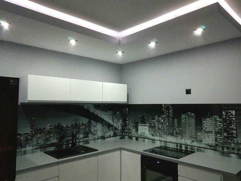 Nowoczesna kuchnia z białymi szafkami, blatem i czarnobiałym panelem szklanym z motywem panoramy miasta podświetlona ledami.