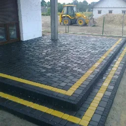 Ułożona kostka brukowa z żółtymi akcentami na schodach wejściowych, widoczny budynek w tle i sprzęt budowlany, świeżo po deszczu.